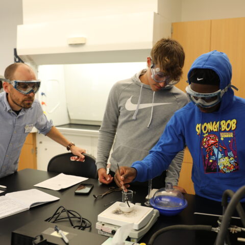 A male teacher watching over two male's chemistry experiment.